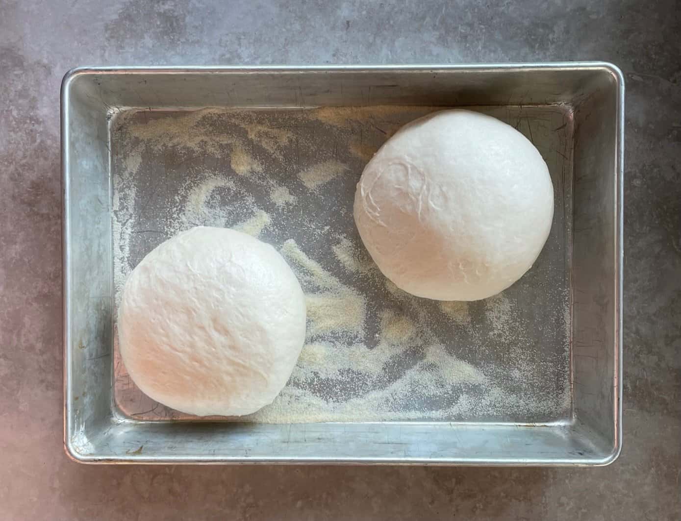 two balls of pizza dough in a baking pan ready for shaping.