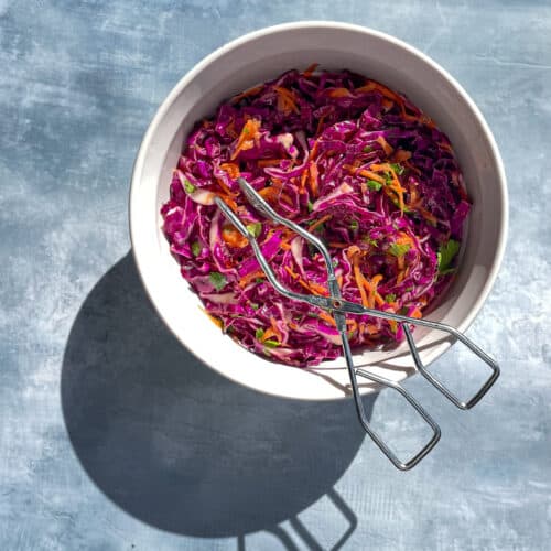 bowl of purple cabbage slaw with shredded carrot, cilantro, and serving tongs in bright sunlight.
