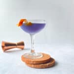 purple cocktail with orange twist in coupe on two stone coasters.