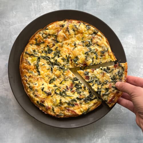 frittata with bacon, kale, and leeks on a plate with a hand removing a cut piece.