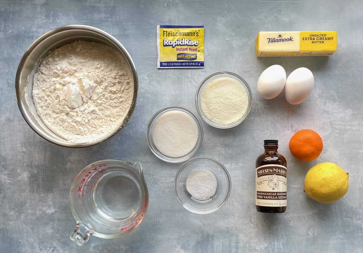 flour, orange, lemon, vanilla, yeast, butter, eggs, sugar, salt, and milk powder on a countertop. 