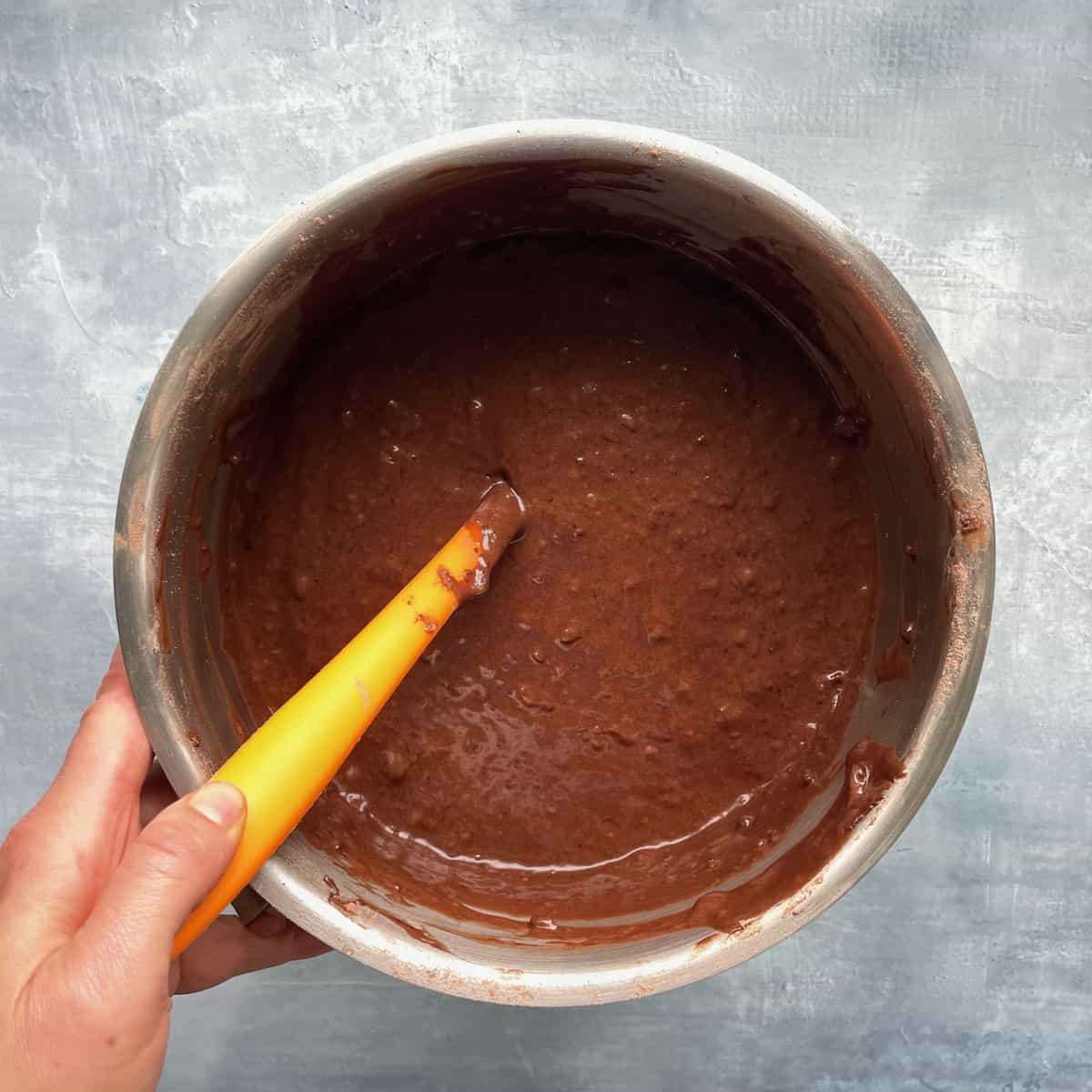 hand holding mixing bowl and orange spatula with the triple chocolate cake batter.