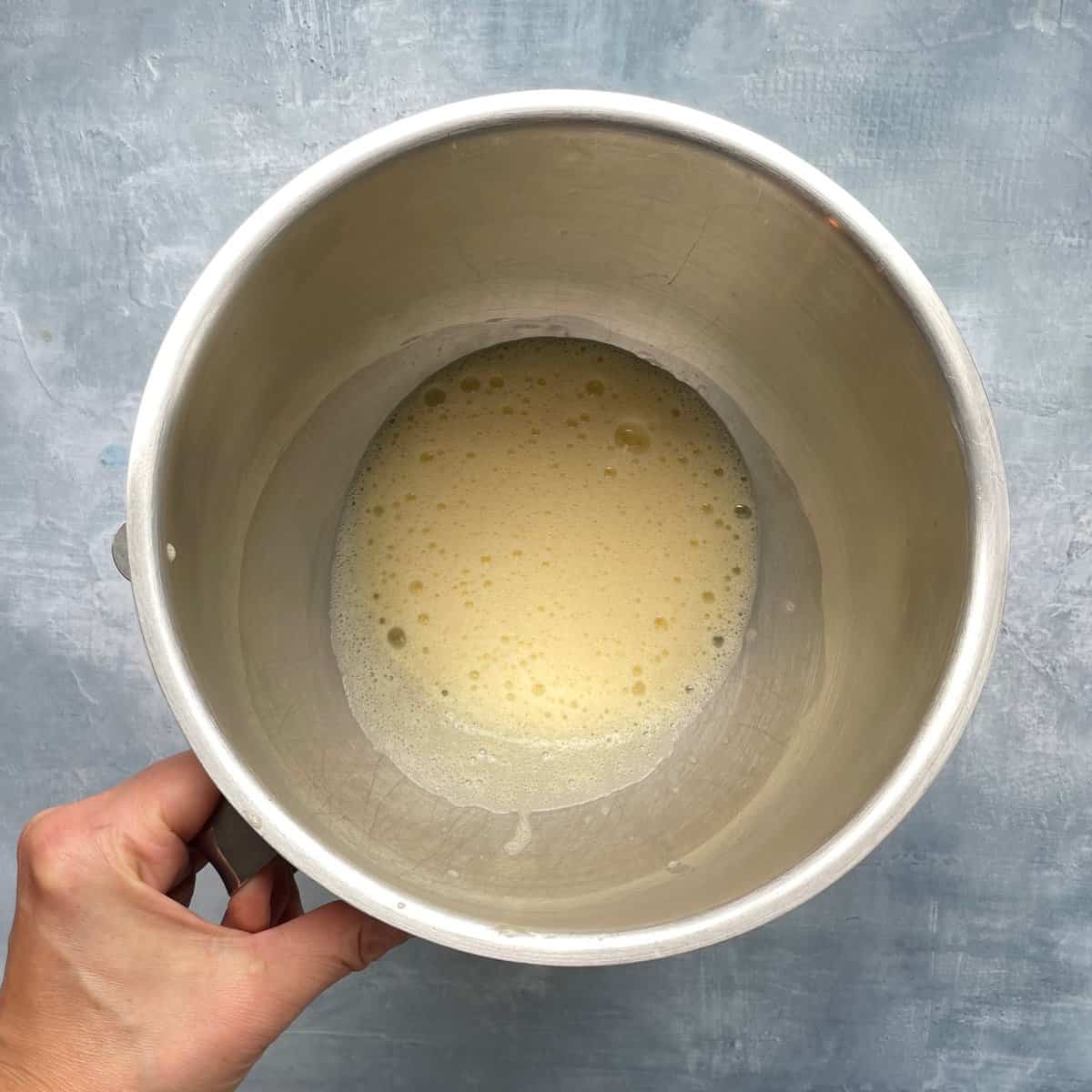 hand holding metal mixing bowl with pale yellow, frothy eggs.