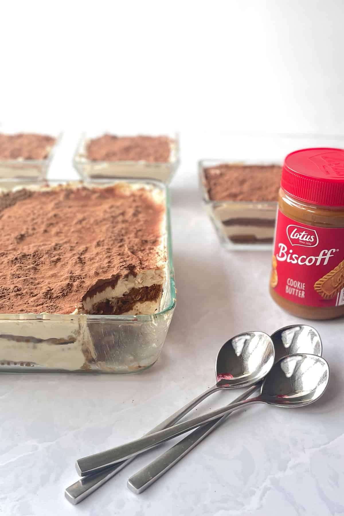 large tiramisu in glass pan with scoop missing next to three spoons and jar of Biscoff cookie butter. 