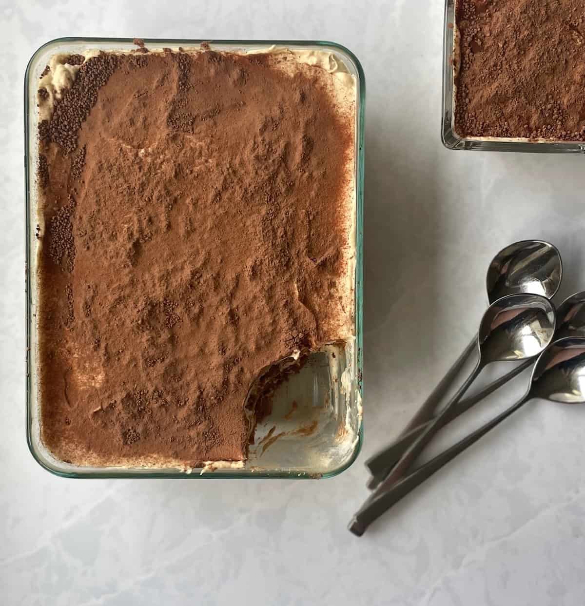 large dish of cocoa-dusted tiramisu minus a scoopful and three spoons next to it.