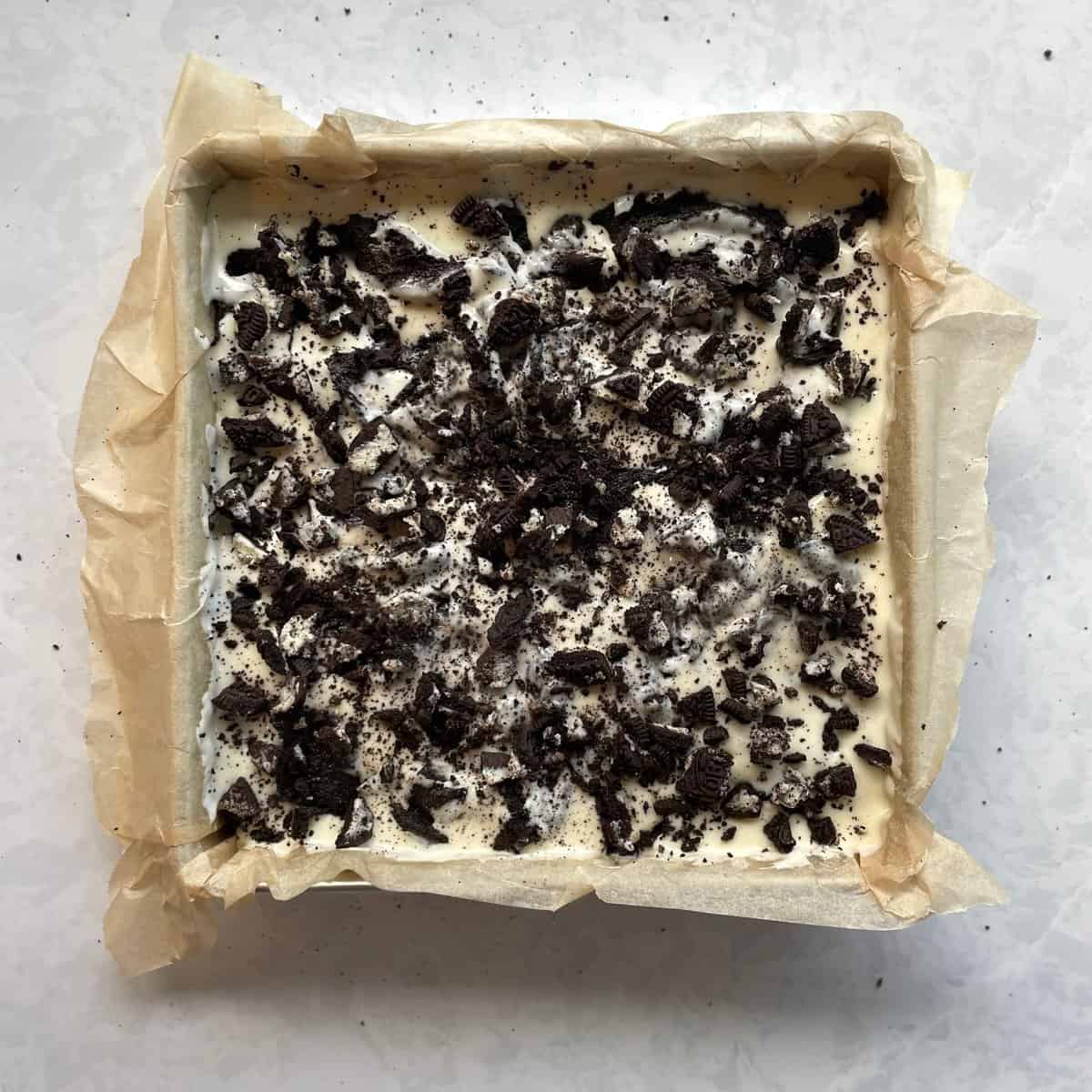 parchment lined square baking pan with chopped Oreos sprinkled atop a cheesecake swirled brownie batter.