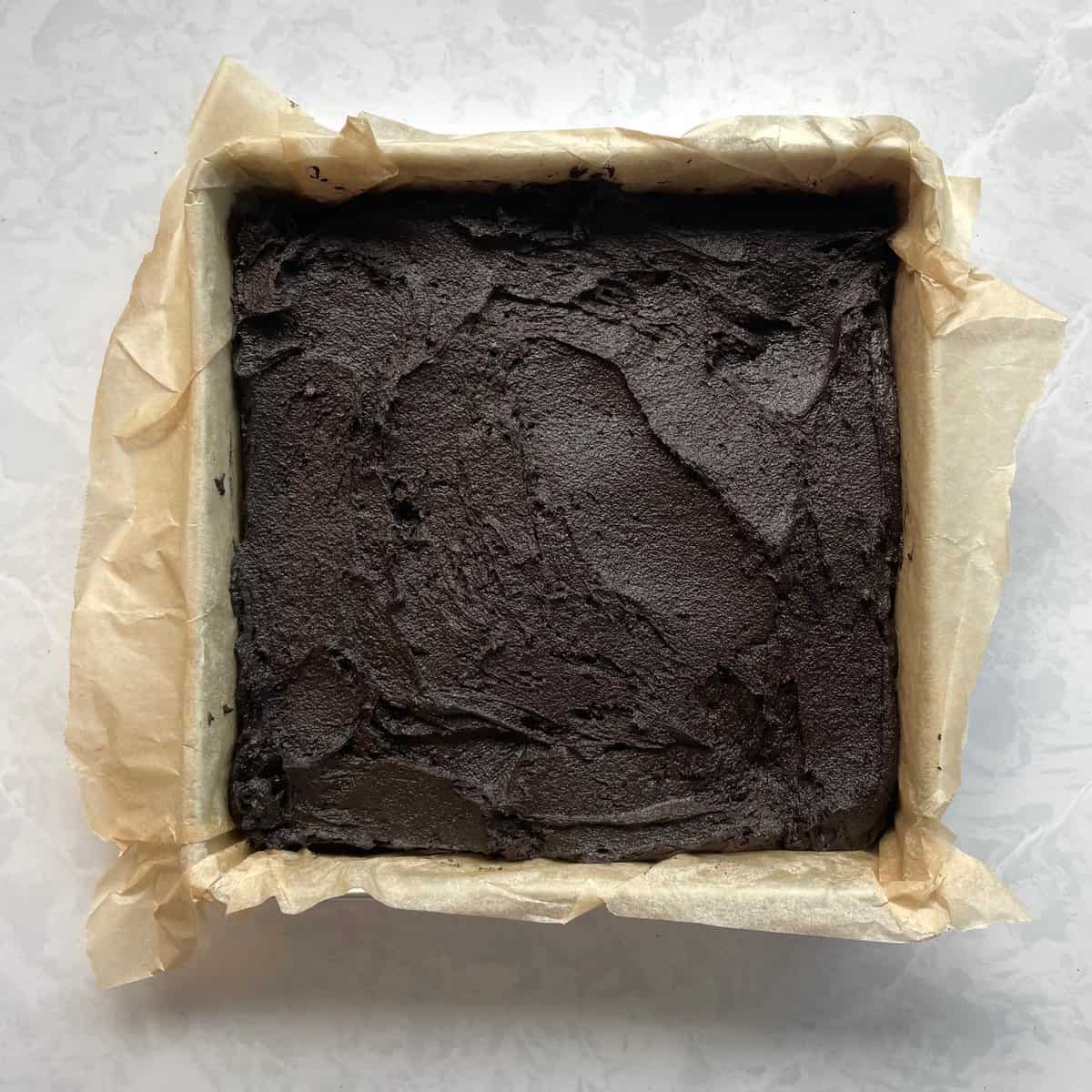 parchment lined square baking pan filled with black brownie batter.