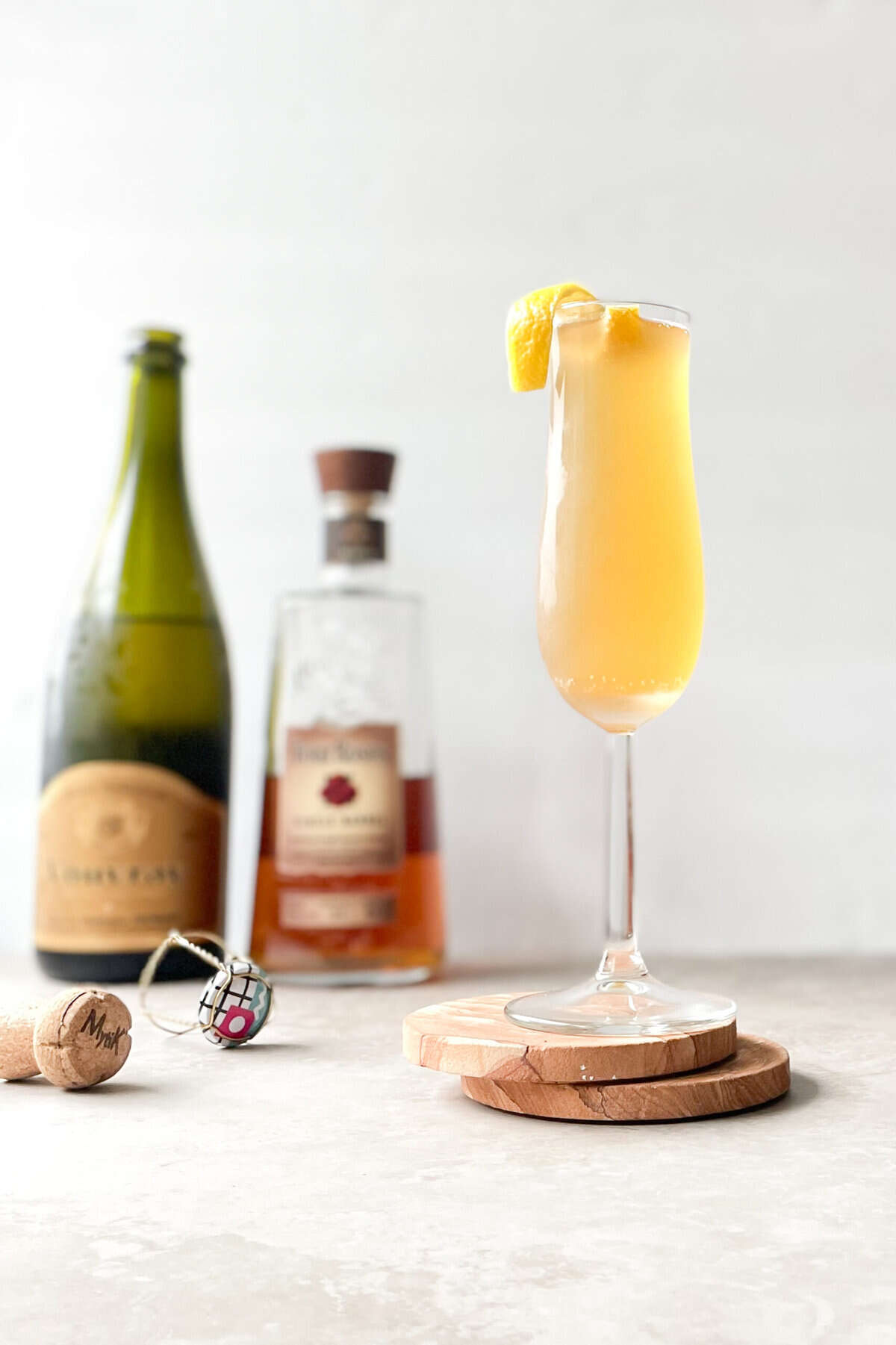 orange cocktail and lemon twist garnish on a champagne flute on two coasters with champagne and whiskey bottle in background.