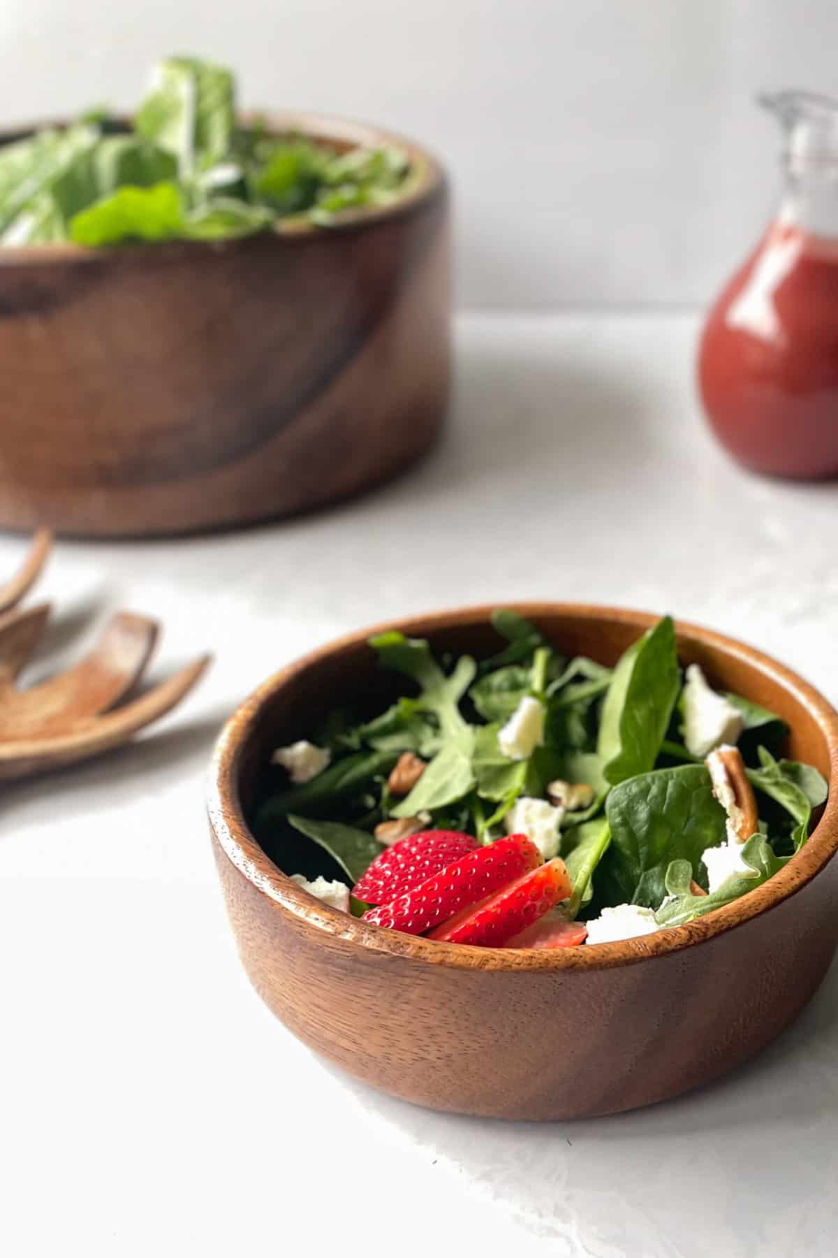 small bowl of arugula spinach salad with strawberries and goat cheese next to salad bowl with greens and tongs and jar of cherry salad dressing.