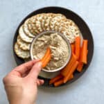 hand with carrot dipping into small bowl of dip on plate with crackers and carrots.