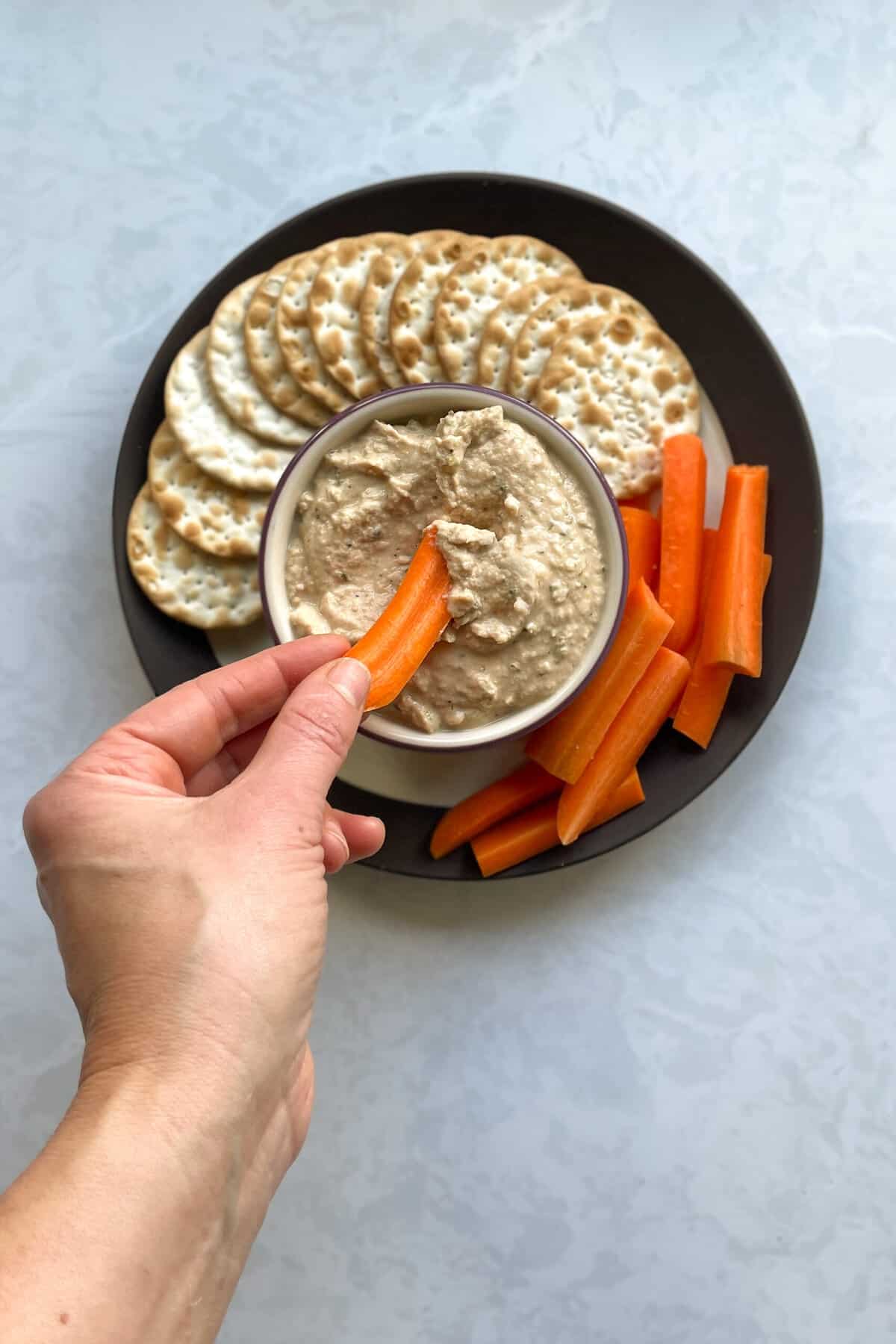 hand dipping a carrot into bowl of dip on plate with crackers and carrots.