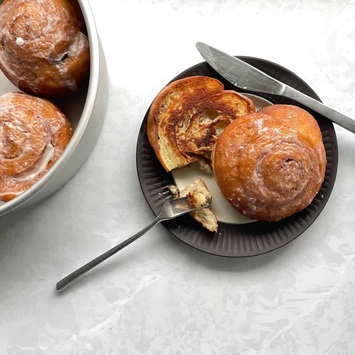 Grilled glazed honey bun on a plate with fork and knife next to pan full of glazed honey buns.