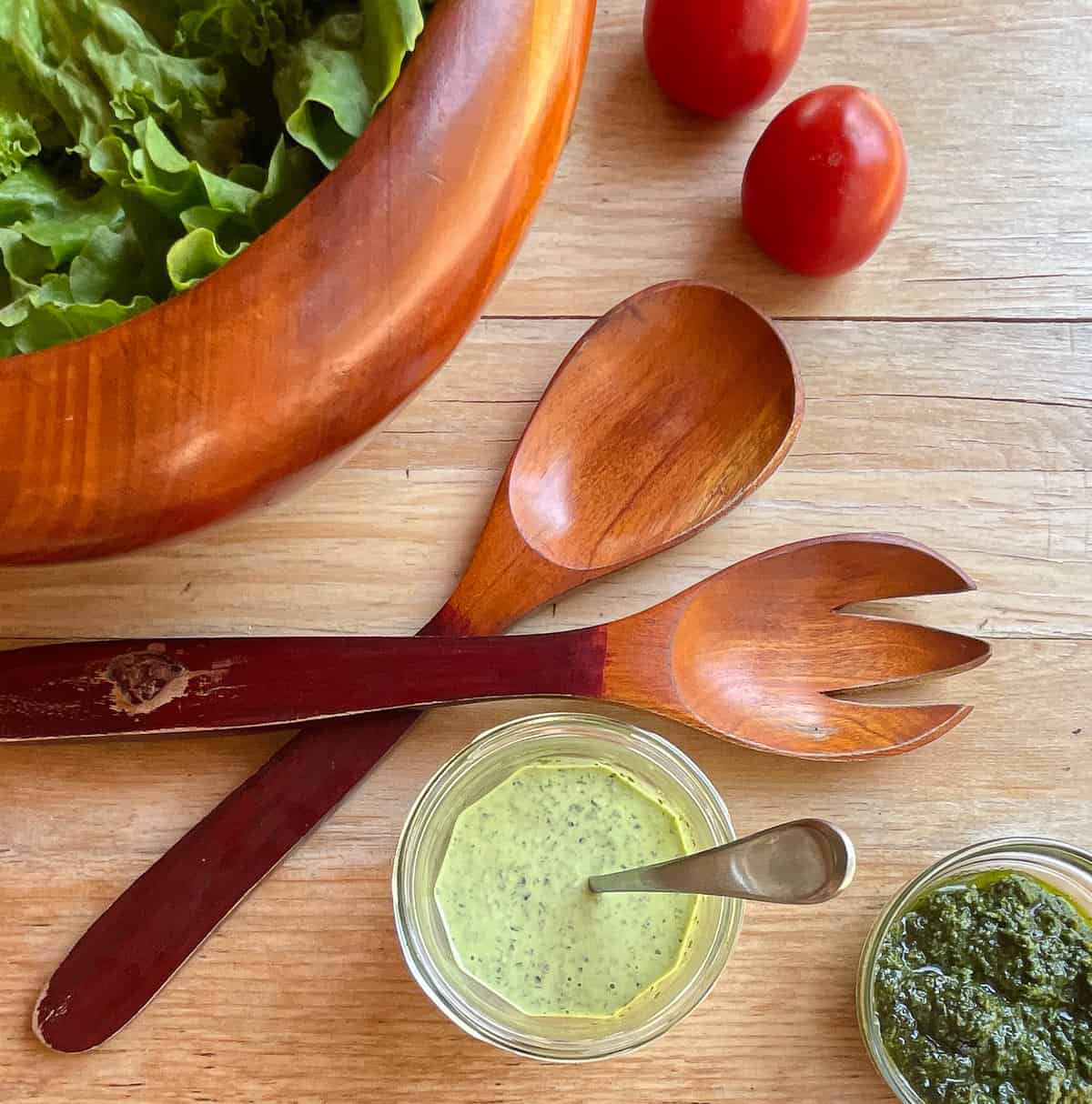 salad bowl, salad tongs, two roma tomatoes, bowl of creamy pesto with spoon, and basil leaves.