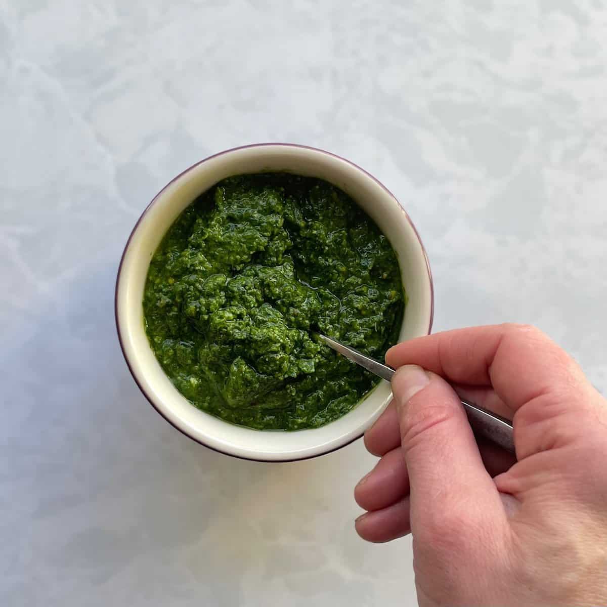 small jar of green pesto with a hand holding a spoon in the pesto.