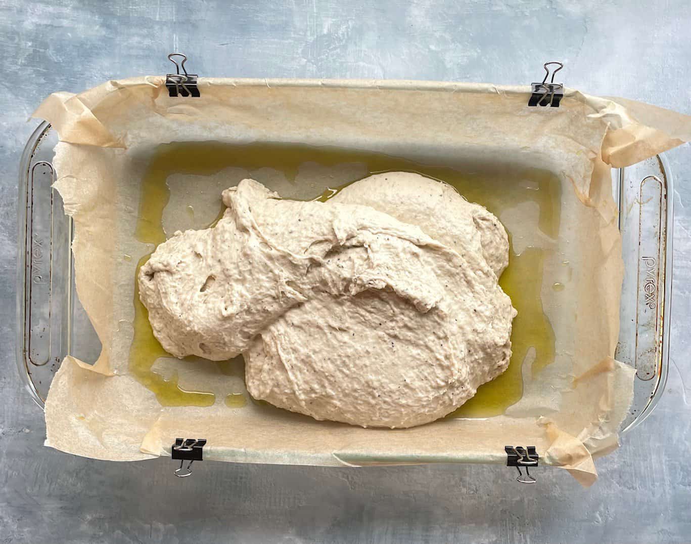 Thin wet focaccia dough in a parchment and olive oil-lined baking dish.