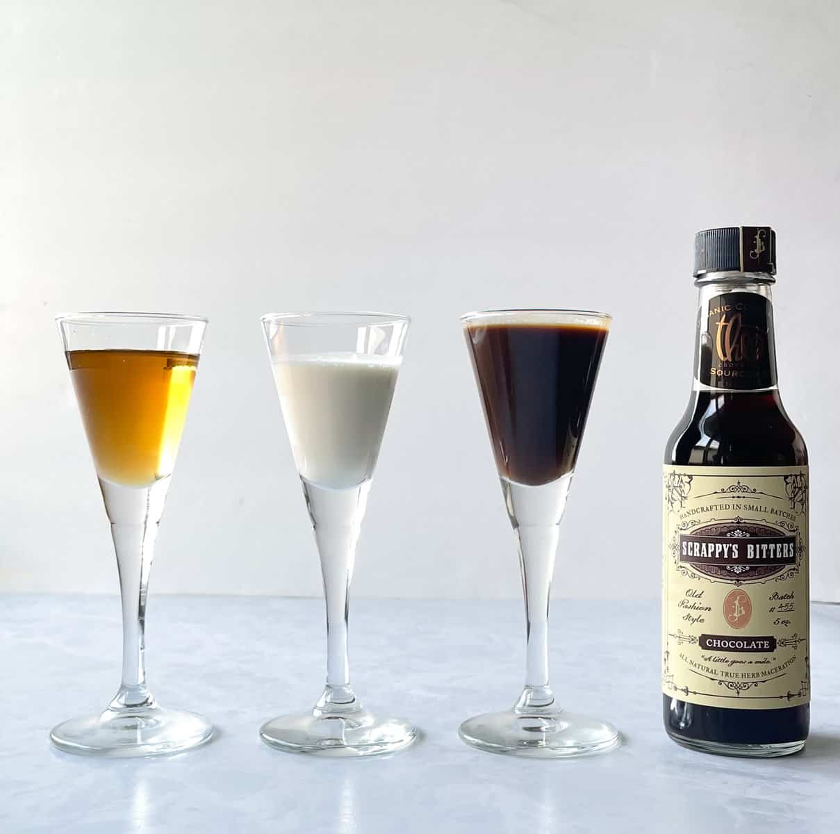 bottle of chocolate bitters and three small glasses of amber, white, and chocolate-colored liquids.