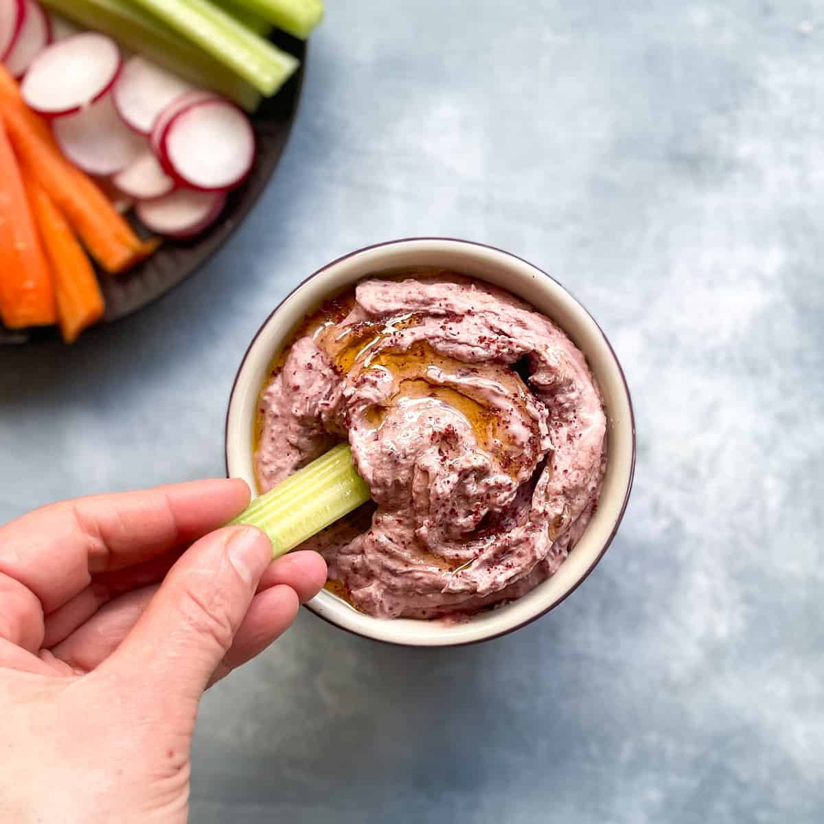 hand holding celery dipping into small bowl of pinkish dip drizzled with olive oil and sumac next to plate of vegetables.
