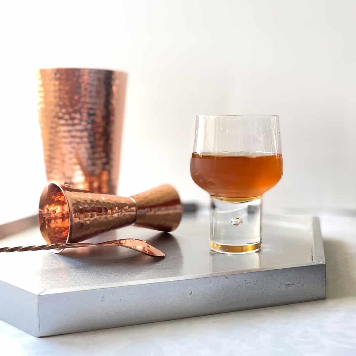 low ball cocktail on a tray with a copper jigger, bar spoon, and cocktail shaker.