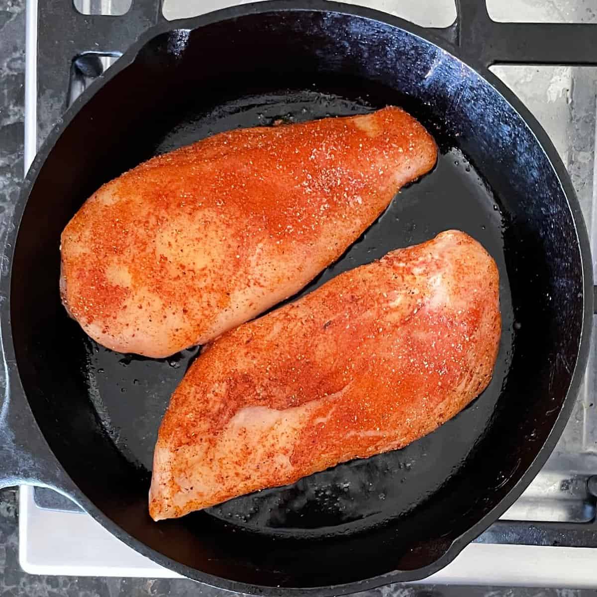two chicken breasts coated in paprika in a cast iron pan on the stove top.