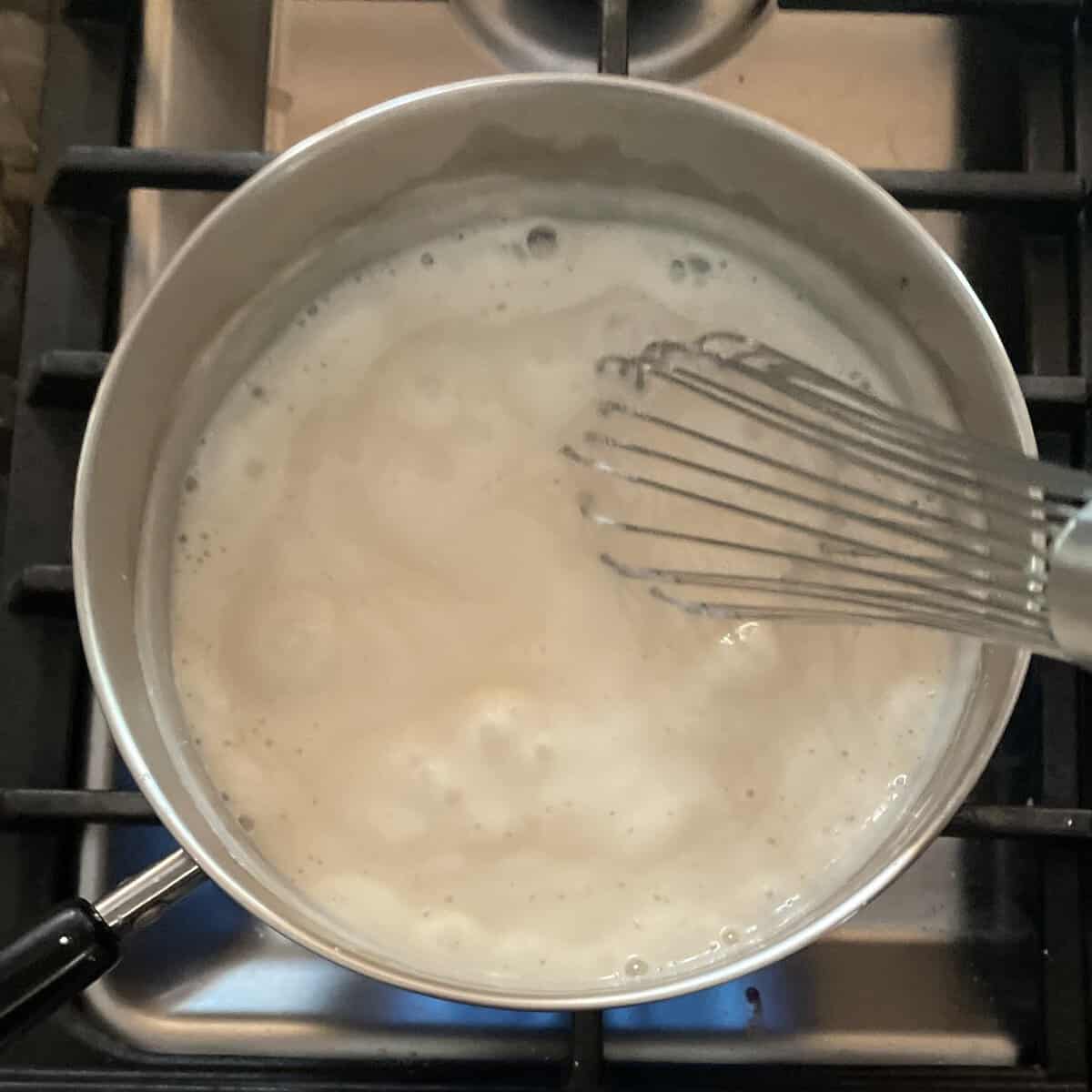 bubbling thick white liquid in a pot on the stove with a whisk in the pot.