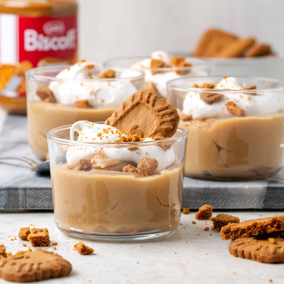 biscoff pudding cups topped with whipped cream and crumbled lotus biscoff cookies. There is a jar of Biscoff cookie butter in the background.