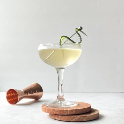 pale yellow cocktail in a coupe on two coasters garnished with a cucumber accordion and next to a brass jigger.