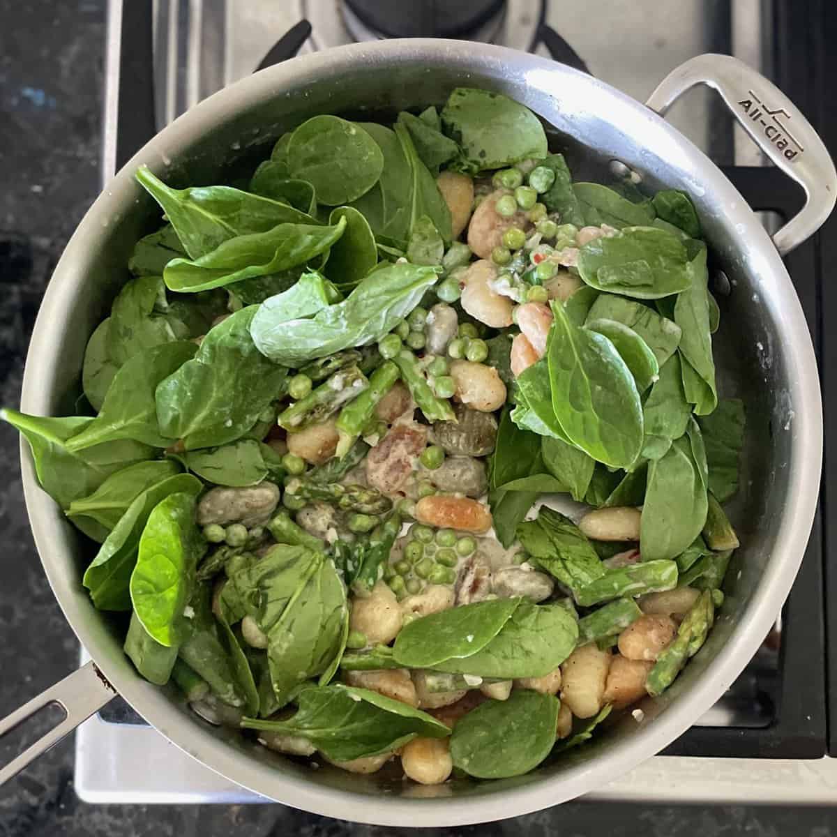 a large metal skillet with fresh spinach leaves added on top of a mixture of peas, asparagus, and gnocchi.