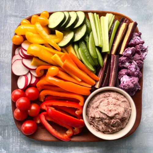 tomatoes, radishes, peppers, carrot sticks cucumber slices, celery, and purple cauliflower florets arranged in rainbow order with a small bowl of mauve colored dip on a square wood tray.