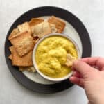 hand dipping a pita chip into a small bowl of bright yellow dip that sits on a plate with pita chips to the side.