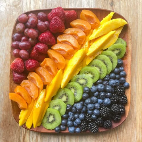 square wood tray with red grapes, strawberries, sliced apricot, sliced mango, sliced kiwi, blueberries, and blackberries in rainbow order.