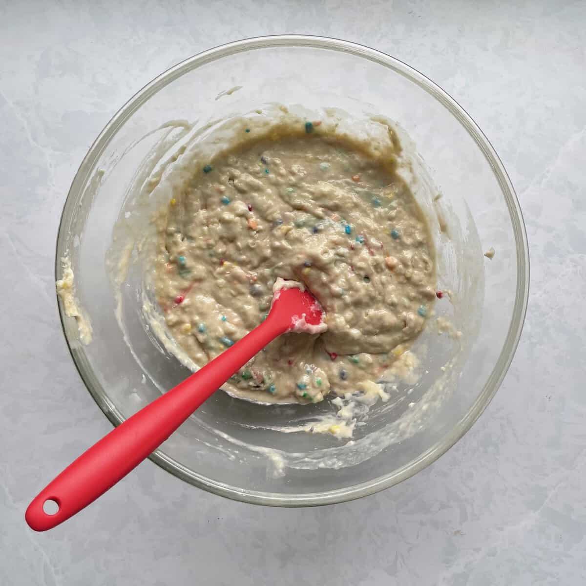 glass bowl with batter with rainbow sprinkles and red silicone spatula.