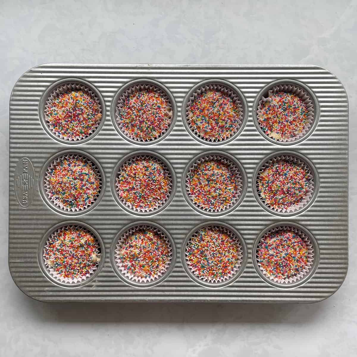 unbaked muffins topped with rainbow nonpareil sprinkles in a muffin tin.