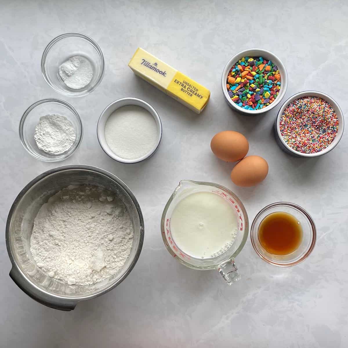 flour, sugar, salt, baking powder and soda, butter, eggs, buttermilk, vanilla, and two types of rainbow sprinkles on a counter.