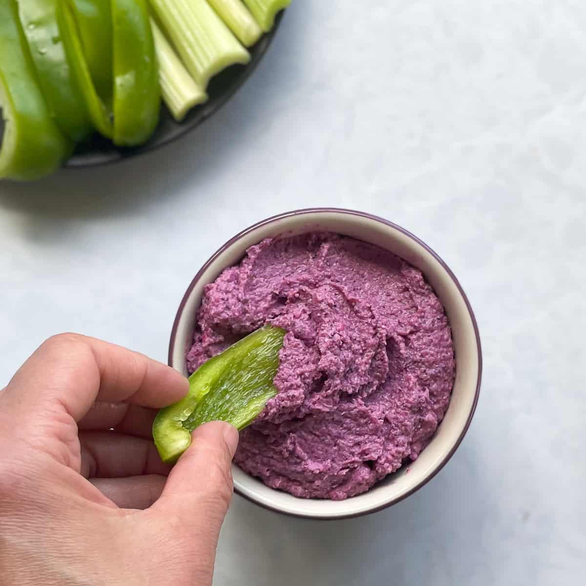bowl of purple dip with hand holding green pepper. There is a plate of green pepper and celery in the corner.