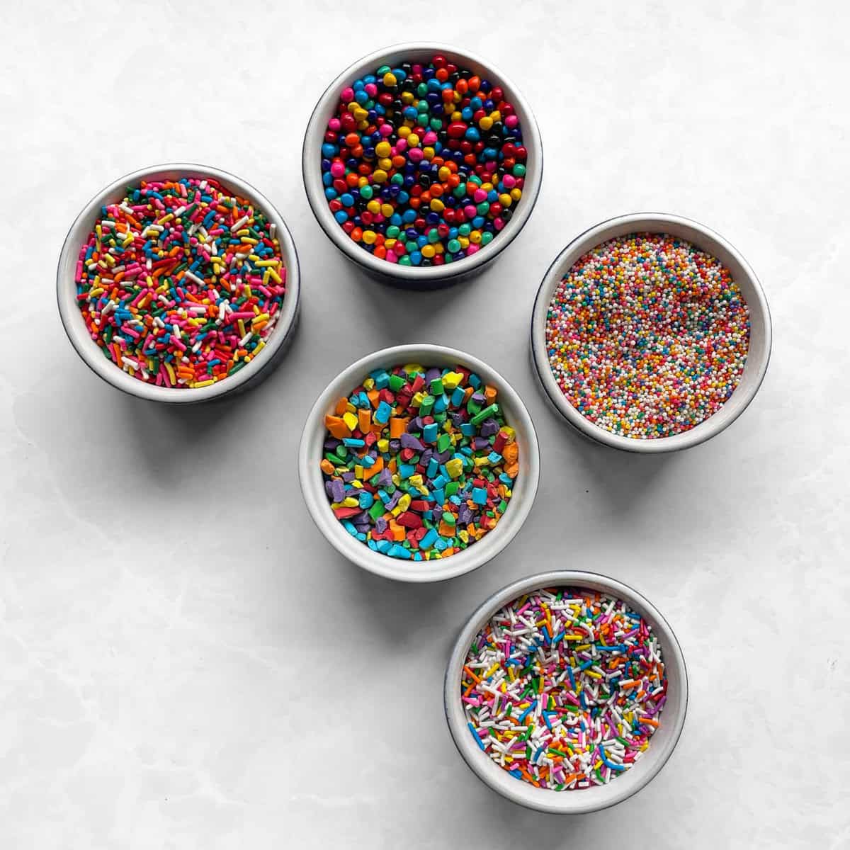 five small bowls of different rainbow sprinkles including rainbow chips and rainbow nonpareils and homemade rainbow sprinkles.