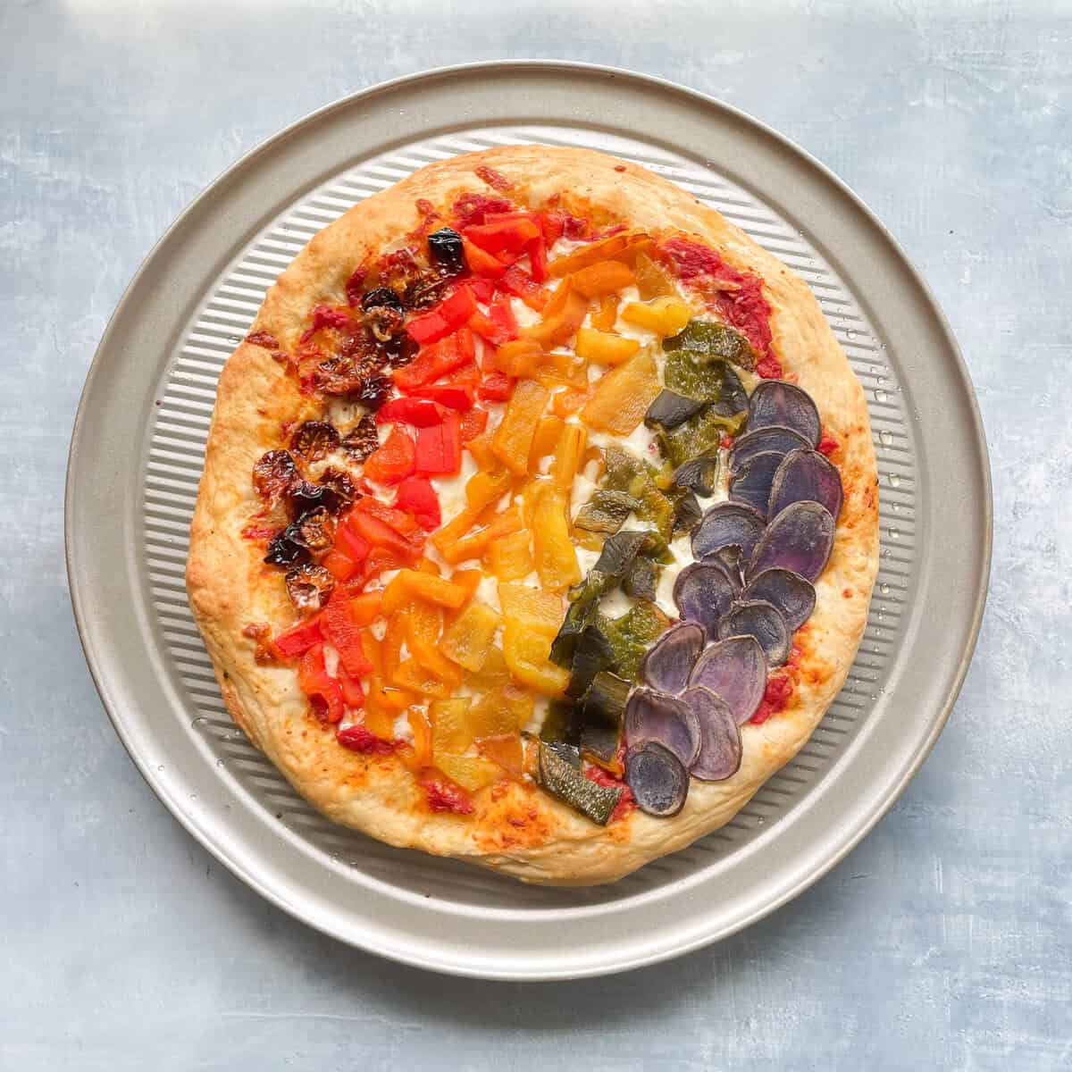 rainbow vegetable pizza on pizza pan with sun-dried tomatoes, bell peppers, poblano pepper, and purple potato slices in rainbow order.