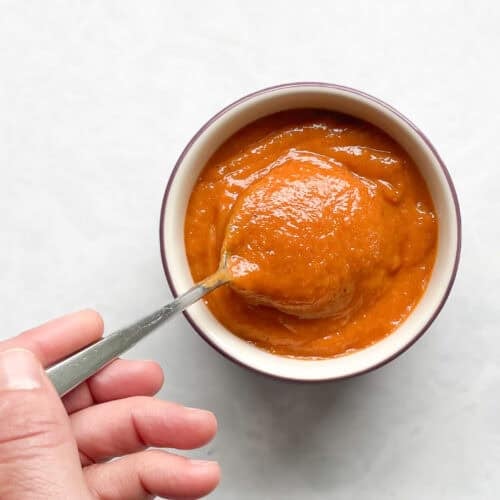 small bowl of bright orange sauce with a hand holding a dipping spoon.
