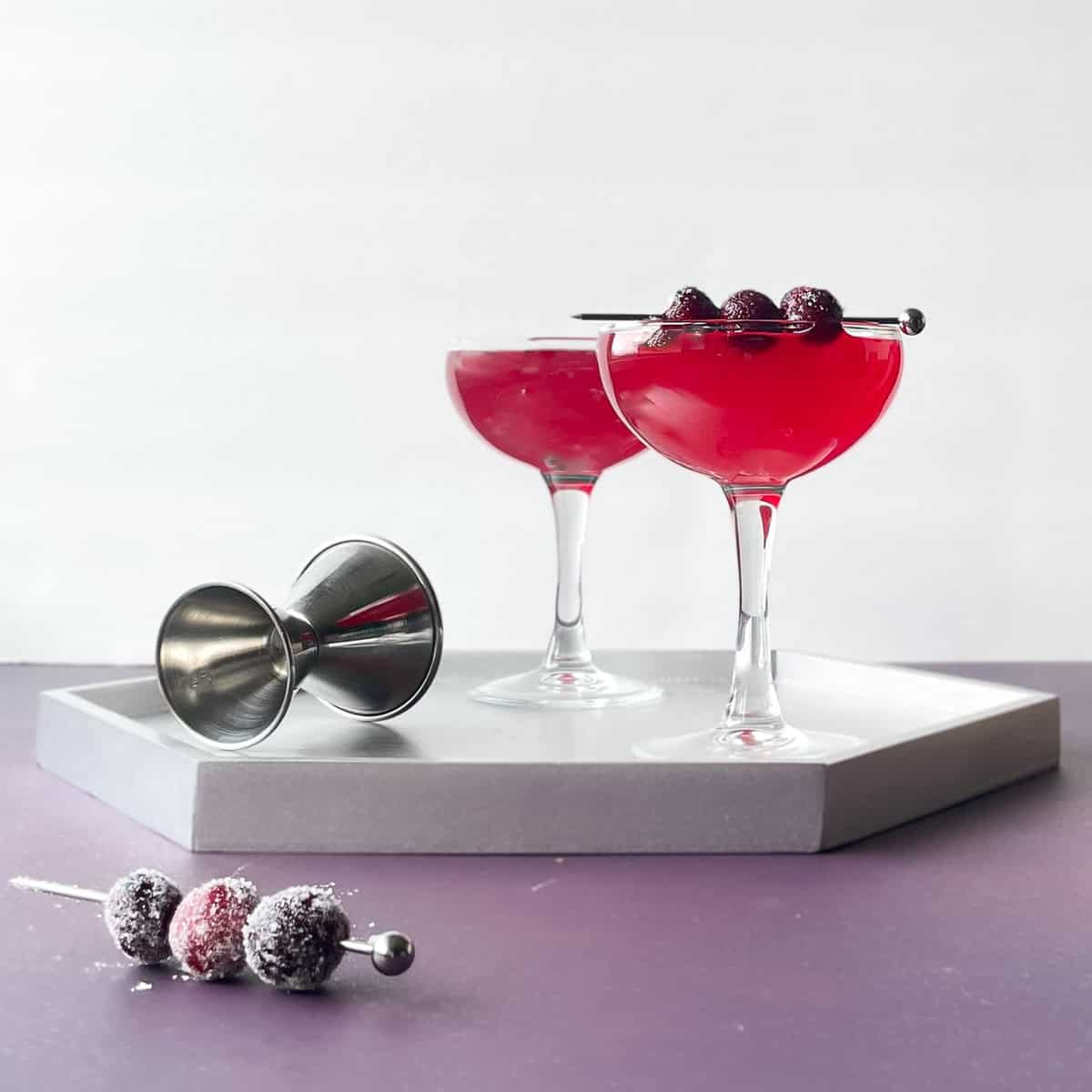 two cranberry gimlets, one garnished with sugared cranberries on a silver tray. There is a silver jigger and pick with three more sugared cranberries.