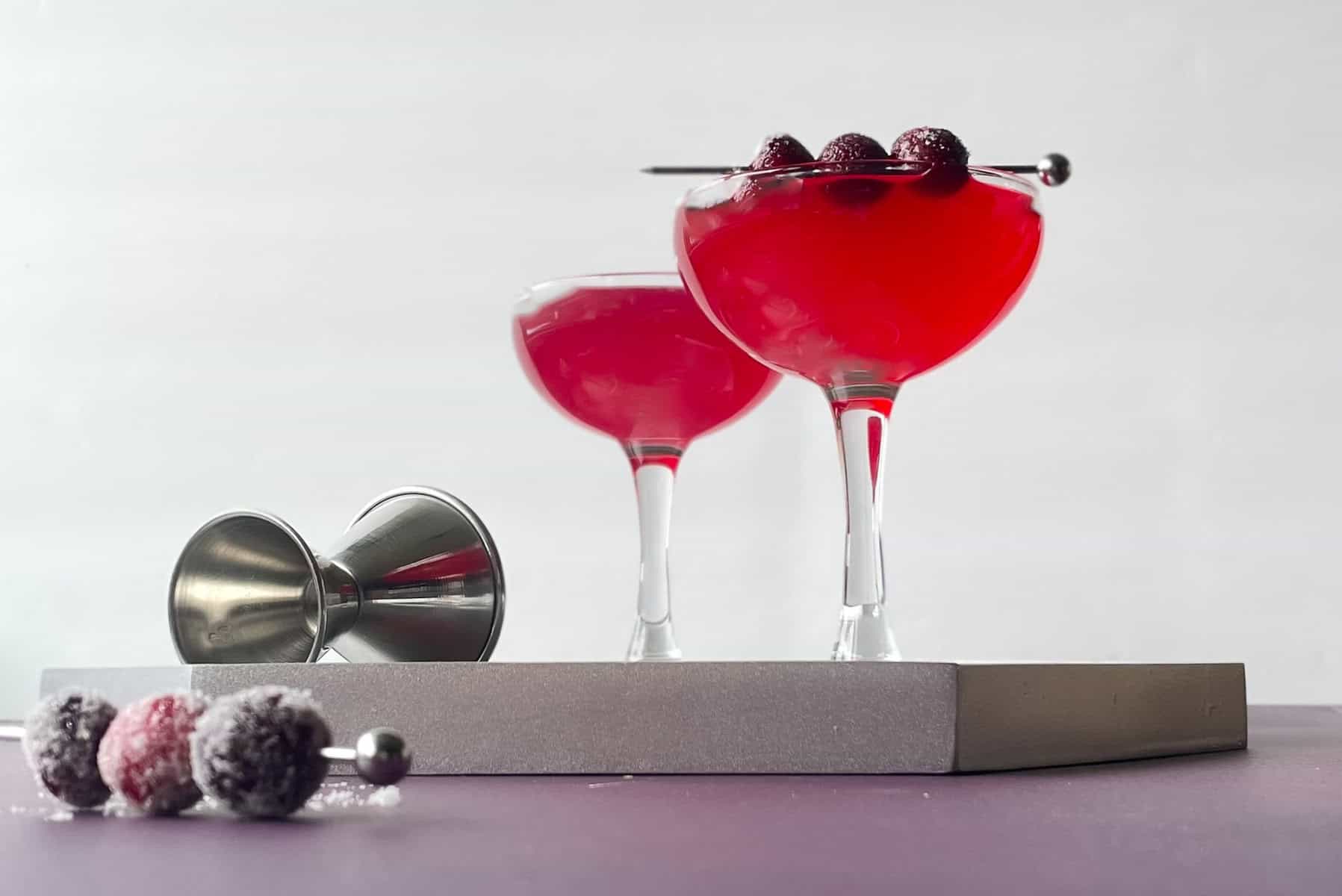 two cranberry cocktails on a silver tray. One is garnished with sugared cranberries. There are more sugared cranberries on a pick and a silver jigger.