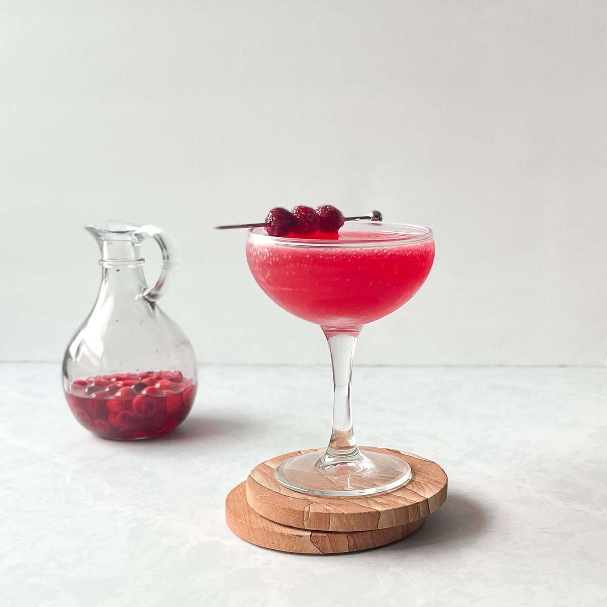 cranberry cocktail in a coupe garnished with sugared cranberries next to a small jug with cranberries in liquid.