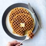 two waffles on a plate with a pat of butter. There is a hand holding a fork with piece of waffle.