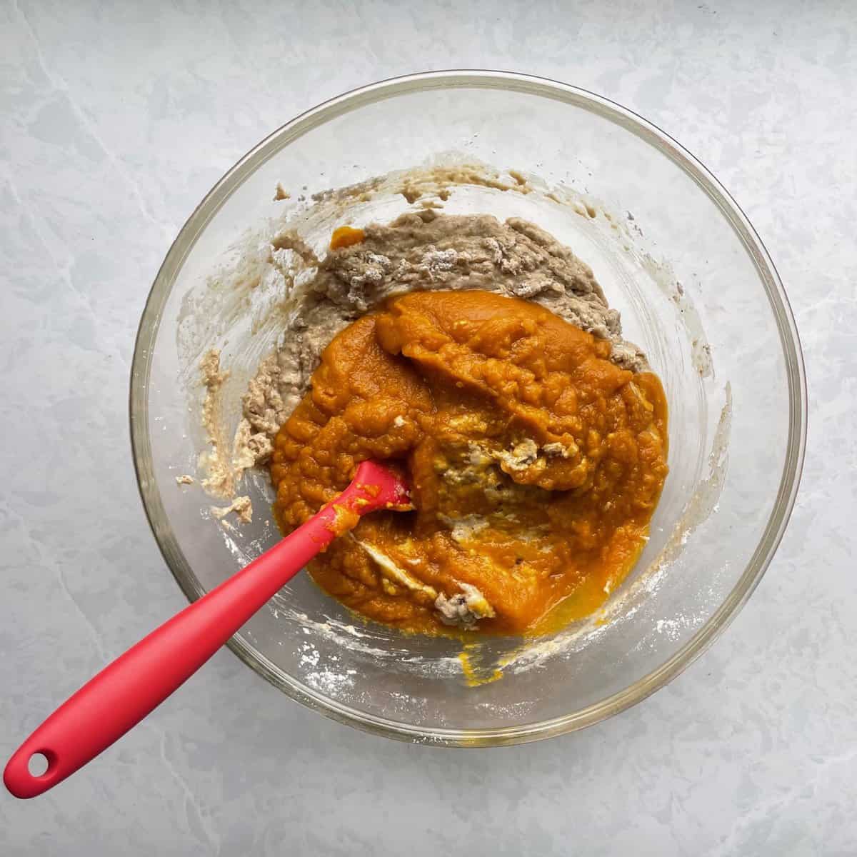 pumpkin puree on top of batter with a red silicone spatula in a glass bowl.