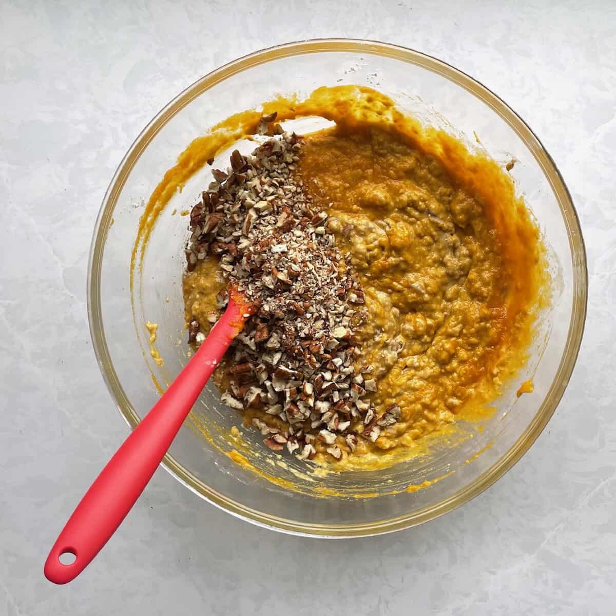 chopped pecans on top of a pumpkin batter with a red spatula in a glass bowl.