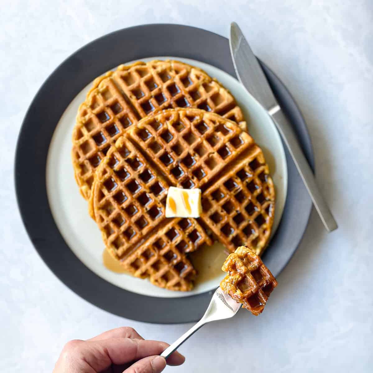  two waffles on a plate with a pat of butter. There is a hand holding a fork with piece of waffle. 