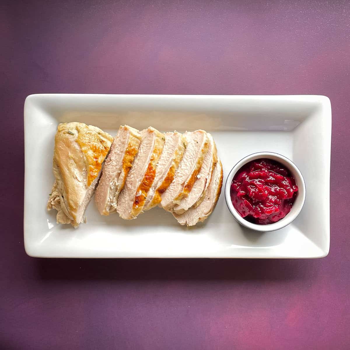 sliced turkey breast with crisp brown skin and small bowl of cranberry sauce on a white tray.