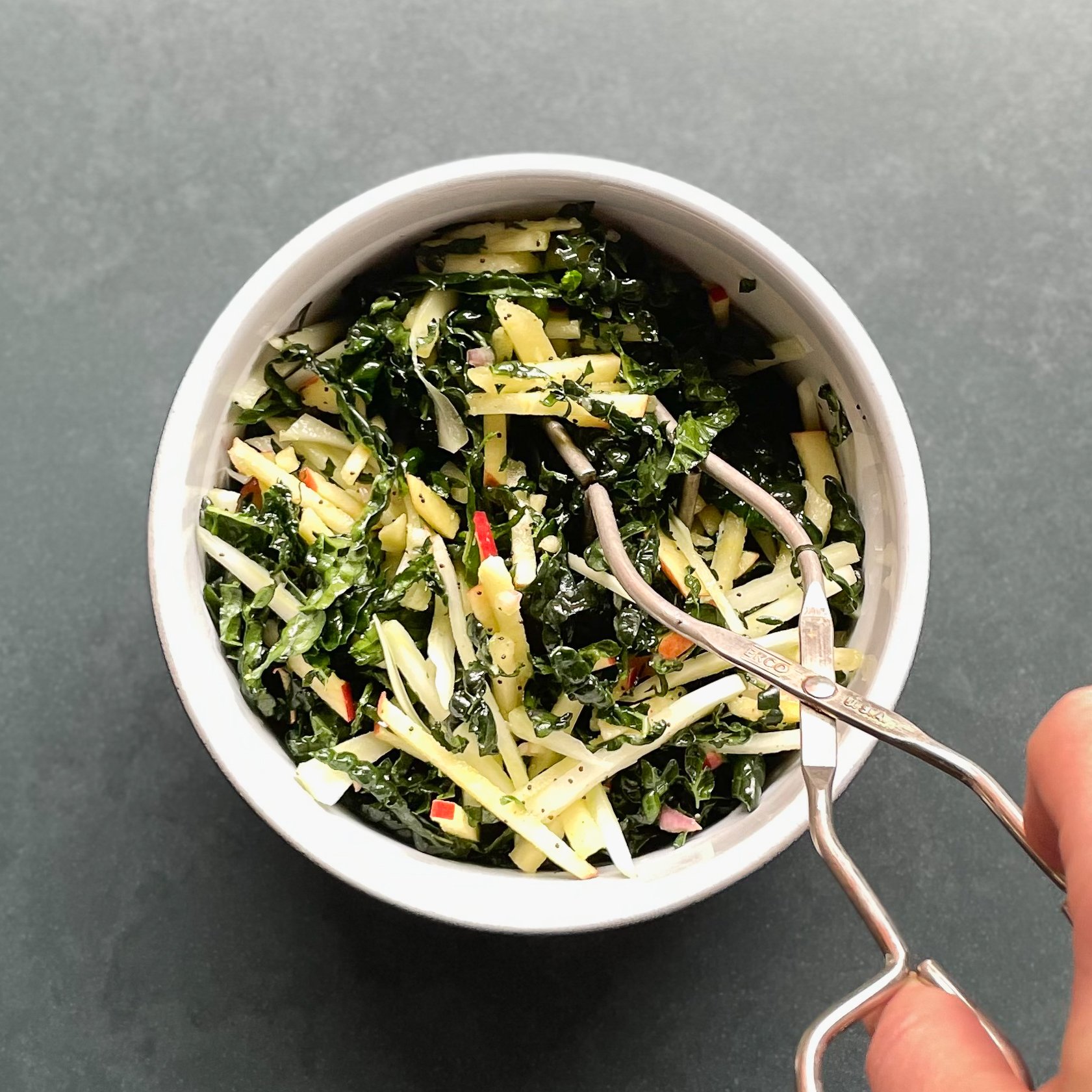 hand holding tongs picking up shredded kale, apple, and fennel from a ceramic bowl.