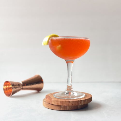 orange cocktail in a coupe garnished with lemon twist on stone coasters next to copper jigger.