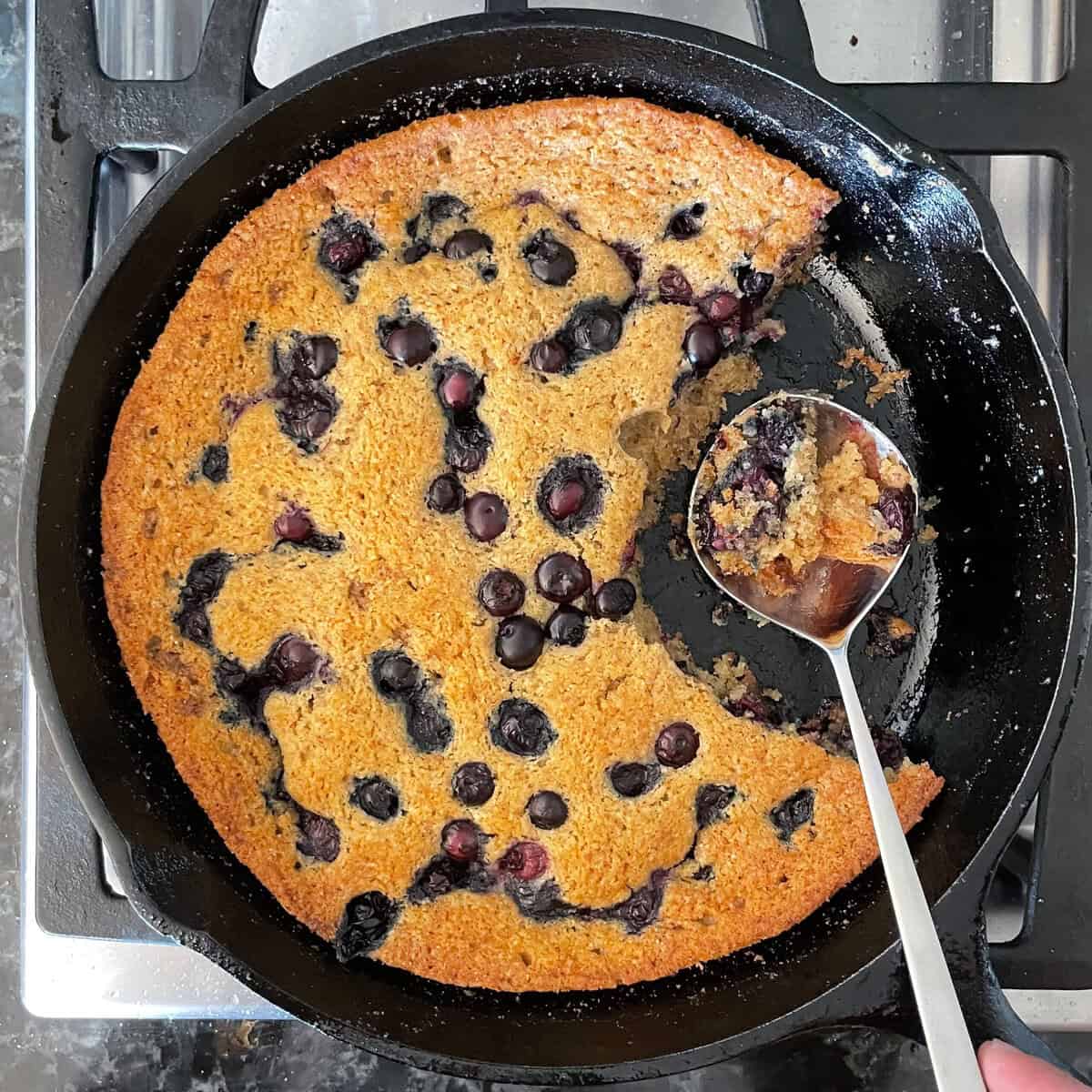 baked blueberry spoon cake in a frying pan with a spoon scooping a bit of blueberry cake.