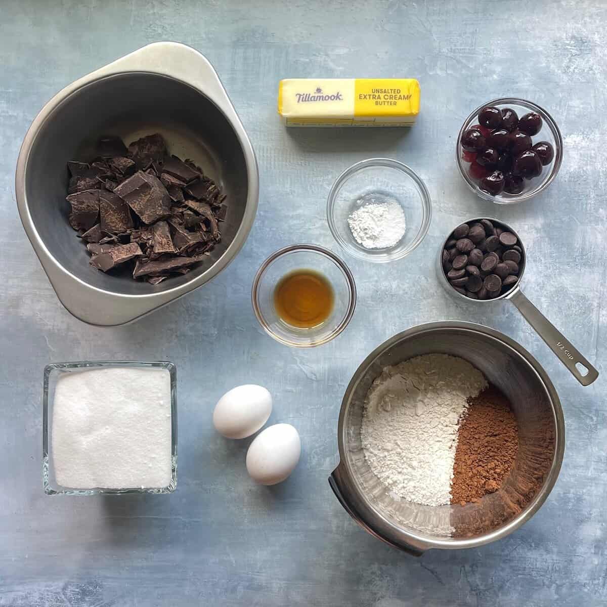 chocolate, cherries, chocolate chips, cocoa, butter, eggs, flour, vanilla, and sugar on a countertop.