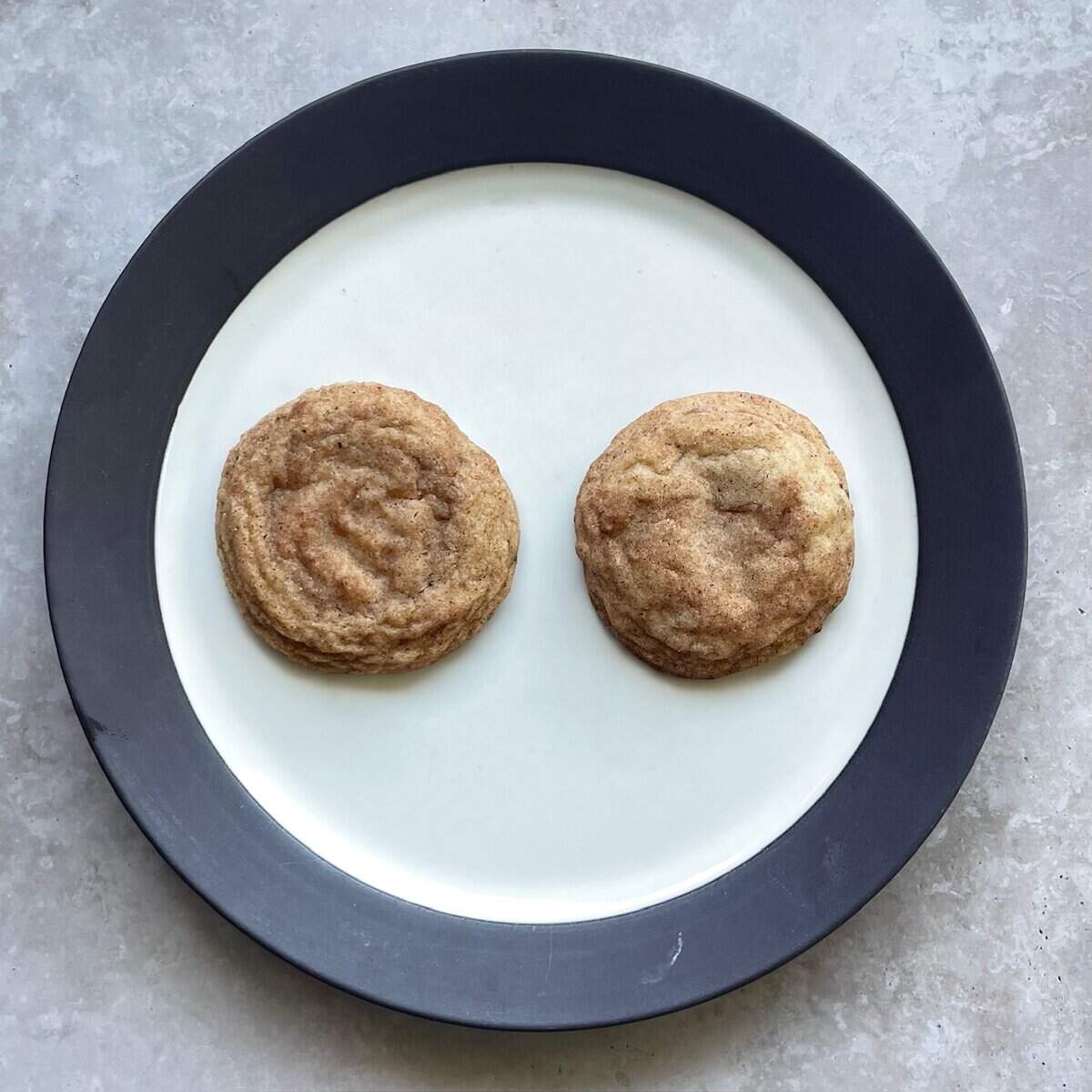 two snickerdoodle cookies on a plate with one thinner and crinkled and the other more puffy.